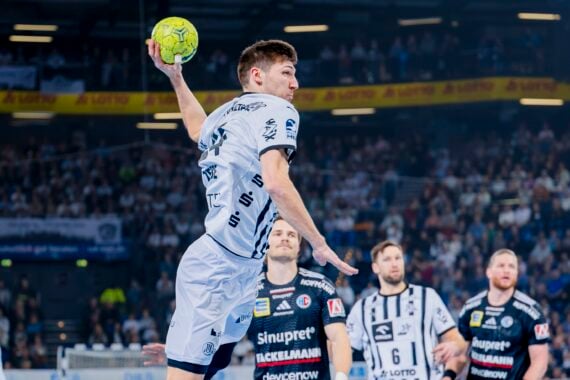 Ein männlicher Handballspieler in einem weiß-blauen Trikot springt in der Luft und bereitet sich darauf vor, einen grünen Ball zu werfen. Er befindet sich auf einem Hallenplatz mit gegnerischen Spielern und einem großen Publikum im Hintergrund.