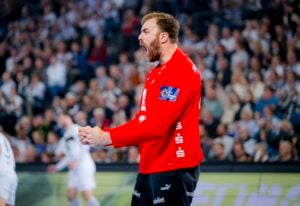 Ein Handballspieler in einem roten Trikot feiert leidenschaftlich auf dem Spielfeld, ballt die Faust und schreit, während im Hintergrund eine verschwommene Menge und andere Spieler zu sehen sind.