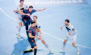 Vier männliche Handballspieler in dunklen und weißen Trikots liefern sich auf einem blauen Hallenplatz ein intensives Spiel. Ein dunkel gekleideter Spieler hält den Ball, während er von zwei weiß gekleideten Spielern eng verteidigt wird; ein weiterer steht in der Nähe und schaut zu.