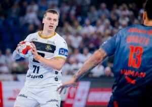 Ein Handballspieler in einem weißen Trikot bereitet sich auf den Wurf vor, während er von einem Gegner in einem blauen Trikot mit der Nummer 13 während eines Hallenspiels verteidigt wird, wobei im Hintergrund Zuschauer zu sehen sind.