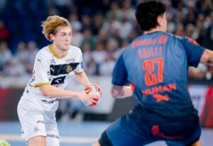 Ein männlicher Handballspieler in einem weißen Trikot bereitet sich darauf vor, den Ball zu werfen, während er von einem Gegner in einem blauen Trikot mit der Nummer 27 verteidigt wird. Die Aktion findet auf einem Hallenplatz mit einer unscharfen Menschenmenge im Hintergrund statt.