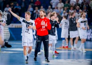 Eine Gruppe männlicher Handballspieler in weißen und schwarzen Trikots feiert auf einem Hallenplatz. Ein Spieler in einem roten Trikot steht in der Mitte, umgeben von klatschenden und jubelnden Mitspielern. Im Hintergrund sind Zuschauer zu sehen.