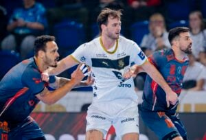 Drei männliche Handballspieler treten während eines Spiels gegeneinander an; der Spieler in Weiß befindet sich zwischen zwei Spielern in Blau und Rot, alle konzentriert und in Bewegung. Die Zuschauer sind im Hintergrund unscharf.