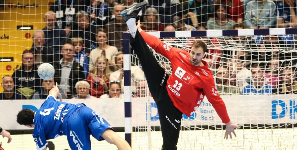 25_nach_TBV_AndiTitel Ein Handballtorwart in einem roten Trikot streckt sein Bein hoch, um einen Schuss abzublocken, während ein Spieler in Blau versucht, ein Tor zu erzielen. Von der Tribüne hinter dem Tor verfolgen die Zuschauer das Geschehen.