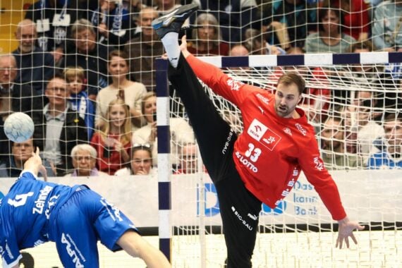 Ein Handballtorwart in einem roten Trikot streckt sein Bein hoch, um einen Schuss abzublocken, während ein Spieler in Blau versucht, ein Tor zu erzielen. Von der Tribüne hinter dem Tor verfolgen die Zuschauer das Geschehen.