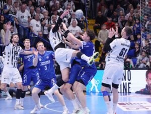 Mehrere Handballspieler in blau-weißen Trikots sind in Aktion, springen und greifen nach dem Ball in einem intensiven Spiel, während im Hintergrund eine große Menschenmenge von der Tribüne aus zusieht.