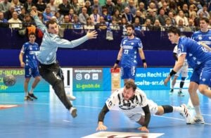 Ein laufendes Handballspiel zeigt einen Spieler in einem weißen Trikot, der auf dem blauen Spielfeld nach vorne springt, während ein Torwart in Grau den Ball abwehrt, während andere Spieler und ein großes Publikum im Hintergrund zusehen.
