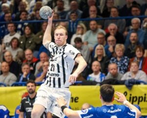 Ein Handballspieler in einem weiß-schwarzen Trikot springt, um den Ball während eines Spiels zu werfen, während im Hintergrund eine Menge von Zuschauern aufmerksam zusieht. Ein anderer Spieler in einem blauen Trikot steht ihm gegenüber.