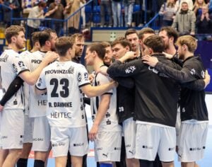Eine Herren-Handballmannschaft in weißen Trikots bereitet sich in einer Halle auf ein Spiel vor. Die Zuschauer schauen von den Tribünen im Hintergrund zu.