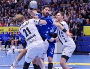 Ein Handballspieler in einem blauen Trikot versucht, ein Tor zu erzielen, während er von zwei Spielern in weißen Trikots während eines intensiven Spiels verteidigt wird, während die Zuschauer im Hintergrund zuschauen.