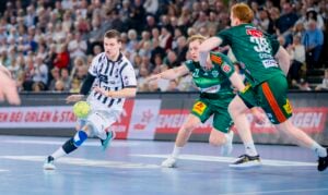 Ein Handballspieler in einem weiß-marine gestreiften Trikot dribbelt den Ball, während zwei Verteidiger in grünen Uniformen versuchen, ihn während eines rasanten Spiels in einer überfüllten Halle zu blocken.