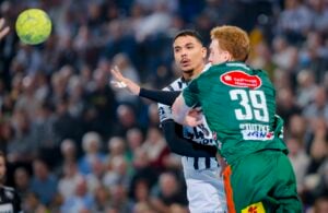 Ein Handballspieler in einem schwarz-weißen Trikot versucht einen Pass zu spielen, während er von einem Gegner in einem grünen Trikot eng verteidigt wird, während eines Spiels mit einer verschwommenen Menschenmenge im Hintergrund.