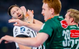 Ein Handballspieler in einem weißen Trikot schaut überrascht, als er während eines Spiels eng mit zwei Spielern in grünen Trikots zusammenspielt. Die Atmosphäre ist intensiv und kämpferisch.