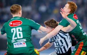 Ein Handballspieler in einem schwarz-weiß gestreiften Trikot versucht, während eines intensiven Spiels zwei Verteidiger in grünen Trikots zu überwinden, wobei ein Verteidiger ihn eng blockiert. Alle Spieler wirken konzentriert und entschlossen.