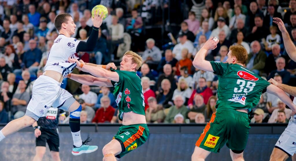 Ein Handballspieler in einem weißen Trikot springt zum Schuss, während zwei Spieler in Grün versuchen, ihn während eines Spiels zu blockieren. Die Zuschauer im Hintergrund schauen gebannt zu.