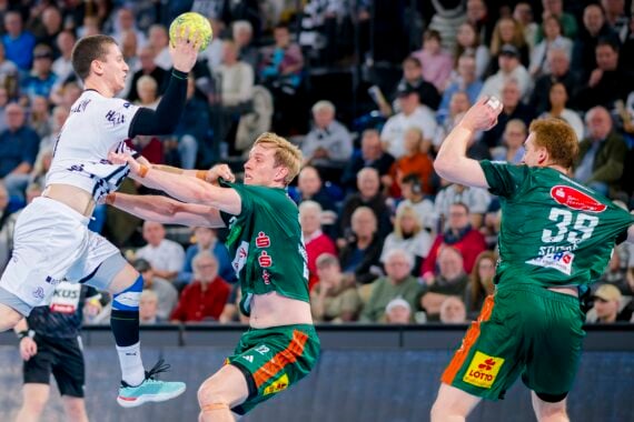 Ein Handballspieler in einem weißen Trikot springt zum Schuss, während zwei Spieler in Grün versuchen, ihn während eines Spiels zu blockieren. Die Zuschauer im Hintergrund schauen gebannt zu.