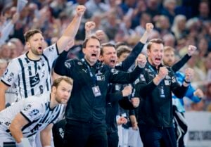 Eine Gruppe von Männern in schwarz-weißen Sportuniformen und -jacken steht zusammen, hebt die Fäuste und schreit aufgeregt, wahrscheinlich beim Feiern eines Handballspiels. Im Hintergrund ist eine jubelnde Menge zu sehen.
