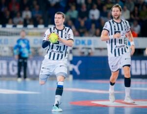 Zwei männliche Handballspieler in weiß-schwarz gestreiften Uniformen laufen auf einem Hallenplatz. Einer hält einen gelben Ball, der andere folgt ihm. Eine Menschenmenge und andere Spieler sind im unscharfen Hintergrund zu sehen.