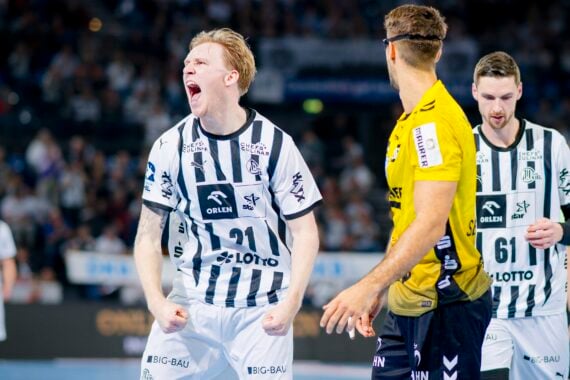 Ein Handballspieler in einem weiß-schwarz gestreiften Trikot jubelt leidenschaftlich und ballt die Fäuste, während ein anderer Spieler in einem gelben Trikot und ein Mannschaftskamerad im Hintergrund während eines Spiels zusehen.