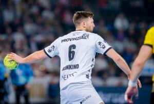 Ein männlicher Handballspieler, der ein weißes Trikot mit der Nummer 6 trägt, bereitet sich während eines Spiels auf den Wurf eines grünen Balls vor. Im Hintergrund sind eine unscharfe Menschenmenge und ein weiterer Spieler in einem gelben Trikot zu sehen.