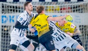 Drei männliche Handballspieler kämpfen um den Ball. Zwei Spieler in weiß-schwarz gestreiften Trikots verteidigen gegen einen Spieler in einem gelb-schwarzen Trikot, der den Ball in Tornähe hält.
