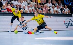 Zwei Spieler in gelben Trikots und ein Spieler in einem schwarz-weiß gestreiften Trikot kämpfen während eines Hallenhandballspiels um einen grünen Ball, während im Hintergrund eine Menge von Zuschauern aufmerksam zusieht.