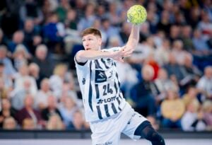 Ein männlicher Handballspieler in einem weiß-schwarz gestreiften Trikot springt mit erhobenem Arm in die Luft, um einen gelben Ball zu werfen, während des Spiels THW Kiel gegen TVB Stuttgart 33:32 Handball-Bundesliga Saison 2025/2026, mit einer unscharfen Zuschauermenge im Hintergrund.