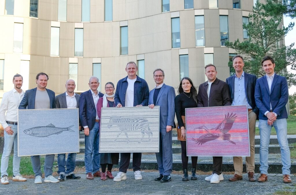 Eine Gruppe von elf Personen steht im Freien vor einem modernen Gebäude. Drei Personen halten ein gerahmtes Kunstwerk, das einen Fisch, ein Zebra und einen Vogel im Flug darstellt.