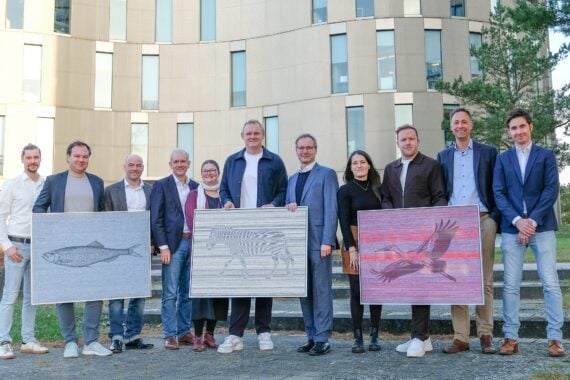 Eine Gruppe von elf Personen steht im Freien vor einem modernen Gebäude. Drei Personen halten ein gerahmtes Kunstwerk, das einen Fisch, ein Zebra und einen Vogel im Flug darstellt.