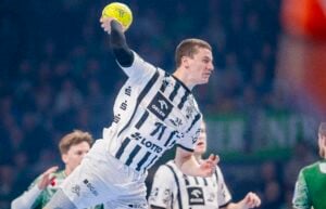 Ein männlicher Handballspieler in einem weiß-schwarzen Trikot springt und bereitet sich darauf vor, während eines Spiels einen gelben Ball zu werfen, mit anderen Spielern und einer unscharfen Menge im Hintergrund.