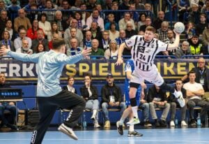 Ein Handballspieler in einem weiß-schwarzen Trikot springt, um den Ball zu werfen, und zielt dabei auf das Tor, während ein Torwart in Hellblau sich darauf vorbereitet, den Ball abzuwehren. Die Zuschauer auf der Tribüne im Hintergrund schauen aufmerksam zu.