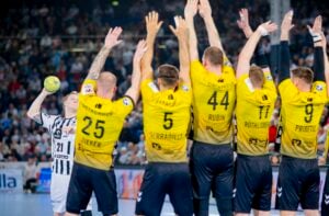Ein Handballspieler in einem schwarz-weiß gestreiften Trikot bereitet sich auf den Wurf vor, während vier Spieler in gelben Trikots mit erhobenen Armen versuchen, ihn während eines Spiels in einer überfüllten Arena zu blockieren.