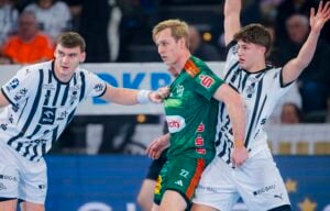 Drei männliche Handballspieler sind während eines Spiels in Aktion. Ein Spieler in einem grünen Trikot steht zwischen zwei Spielern in weiß-schwarz gestreiften Trikots. Die Spieler sind konzentriert, einer hebt die Arme zum Blocken.