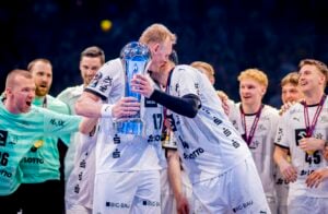 Eine Gruppe männlicher Handballspieler in weißen Trikots feiert auf dem Spielfeld. Zwei Spieler in der Mitte küssen eine große blaue Trophäe, während die Mannschaftskameraden um sie herum jubeln und lächeln. Die Atmosphäre ist freudig und triumphal.