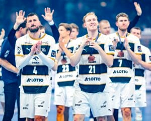 Eine Gruppe männlicher Handballspieler in weiß-goldenen Trikots steht zusammen auf einem Spielfeld, klatscht und lächelt, einige heben die Hände im Hintergrund. Die Stimmung scheint feierlich oder anerkennend zu sein.