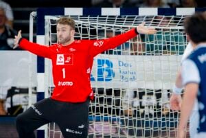 Ein Handballtorwart in einem roten Trikot steht mit ausgestreckten Armen vor dem Tor und bereitet sich darauf vor, einen Schuss abzufangen. Im Vordergrund sind Spieler in Blau und Weiß zu sehen.