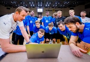 Eine Gruppe von männlichen Sportlern in blauen Trikots sitzt dicht gedrängt um einen Tisch und beobachtet aufmerksam einen Trainer in Weiß, der in einer Sporthalle etwas auf einem Laptop erklärt.