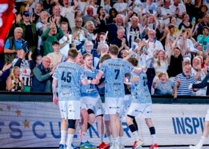 Fünf männliche Handballspieler in blau-weißen Trikots feiern gemeinsam auf dem Spielfeld, während im Hintergrund eine große, jubelnde Menge steht und begeistert applaudiert.