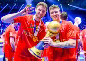 Zwei lächelnde männliche Athleten in roten Trikots feiern und halten eine goldene Trophäe mit blauem Sockel, auf der IHF steht. Einer blinzelt mit drei Fingern; beide tragen Medaillen um den Hals auf einem hell erleuchteten Platz in einer Sportarena. Im Hintergrund sind Mannschaftskameraden zu sehen.