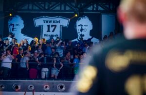 Eine Menschenmenge sitzt und steht in einer Arena unter einem großen Transparent mit der Nummer 17, dem Namen Wiencek und zwei Schwarz-Weiß-Porträts. Der Vordergrund ist unscharf und zeigt den Rücken einer Person, die ein schwarzes Hemd trägt.