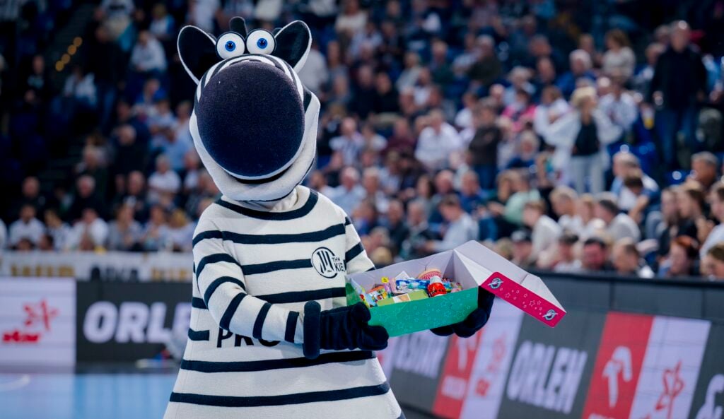 Eine Person in einem Zebra-Maskottchen-Kostüm hält eine offene Schachtel mit bunten Gegenständen in der Hand und steht auf einem Sportplatz mit einer unscharfen Menschenmenge im Hintergrund.