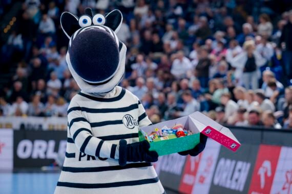 Eine Person in einem Zebra-Maskottchen-Kostüm hält eine offene Schachtel mit bunten Gegenständen in der Hand und steht auf einem Sportplatz mit einer unscharfen Menschenmenge im Hintergrund.