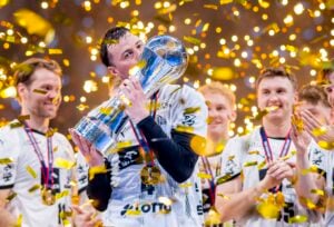 Ein jubelnder Sportler in einem weißen Trikot küsst eine große silberne Trophäe, umgeben von jubelnden Mannschaftskameraden und goldenem Konfetti. An ihren Hälsen hängen Medaillen, die den Sieg einer Meisterschaft feiern.