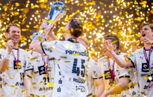 Eine Sportmannschaft in weißen Trikots feiert, während ein Spieler eine Trophäe hochhebt. Um sie herum fällt goldenes Konfetti, und ihre Gesichter zeigen Aufregung und Freude.