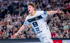 Ein Handballspieler in einem weiß-blauen Trikot mit der Nummer 22 jubelt mit ausgestreckten Armen während eines Spiels, während im Hintergrund eine jubelnde Menge zu sehen ist.