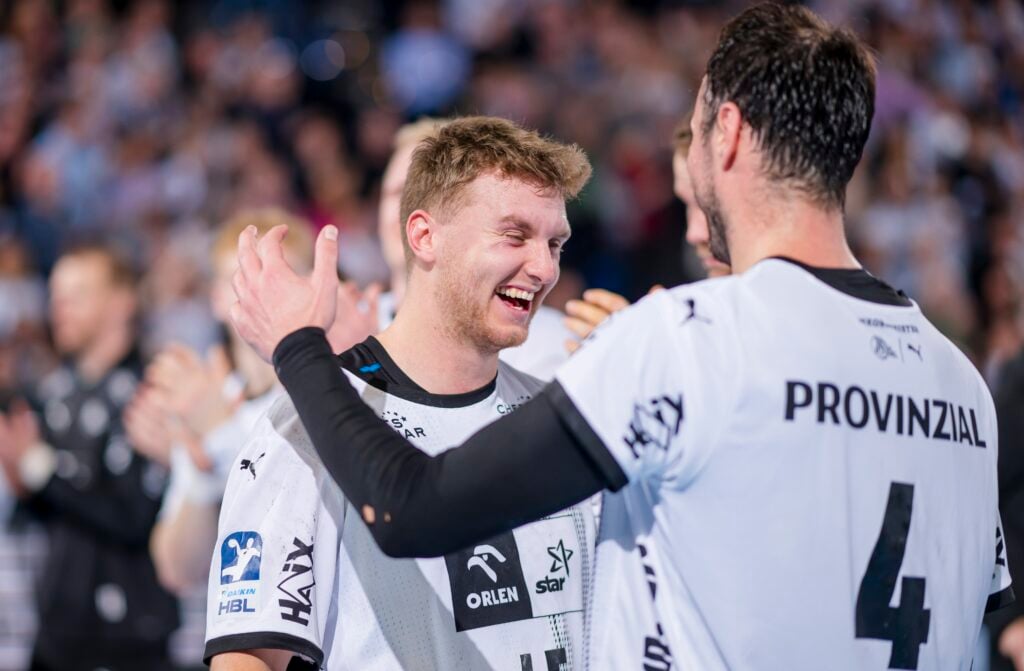 Zwei männliche Athleten in weißen Trikots lächeln und feiern gemeinsam auf einem Sportplatz, wobei der eine seine Hand auf die Schulter des anderen legt. Im Hintergrund sind unscharfe Zuschauer zu sehen.