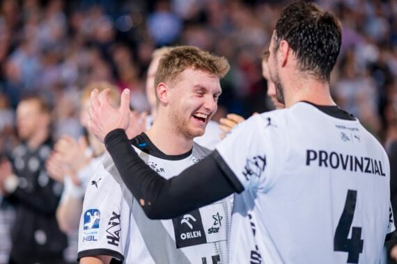 Zwei männliche Athleten in weißen Trikots lächeln und feiern gemeinsam auf einem Sportplatz, wobei der eine seine Hand auf die Schulter des anderen legt. Im Hintergrund sind unscharfe Zuschauer zu sehen.