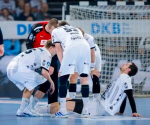 Ein Handballspieler sitzt auf dem Spielfeld und hält sich das Bein vor Schmerzen, während Mannschaftskameraden und ein Schiedsrichter nach ihm sehen. Im Hintergrund sind ein Tor und Zuschauer in einer Halle zu sehen.