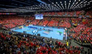 Eine große Halle voller jubelnder Fans in Rot, die zwei Handballmannschaften beobachten, die sich auf dem blauen Spielfeld für eine Zeremonie aufstellen, bei der Fahnen und Offizielle anwesend sind. Eine große Leinwand zeigt die Szene über dem Kopf.