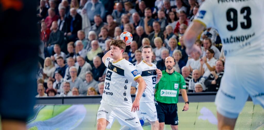 Ein Handballspieler in einem weißen Trikot bereitet sich darauf vor, den Ball während eines Spiels zu fangen. Zu sehen sind Mannschaftskameraden, ein Gegner und ein Schiedsrichter; im Hintergrund sieht eine Zuschauermenge zu.